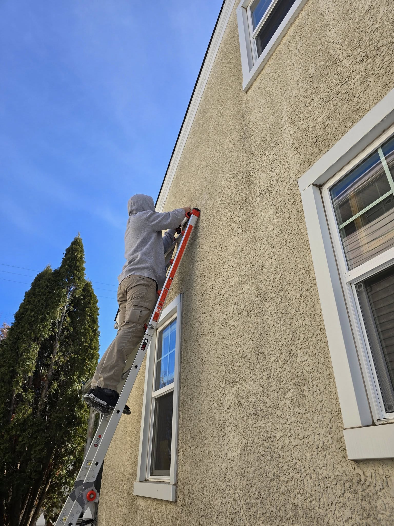 Person wearing a gray hoodie and beige pants standing on an extension ladder working on the side of a beige stucco house under a clear blue sky.